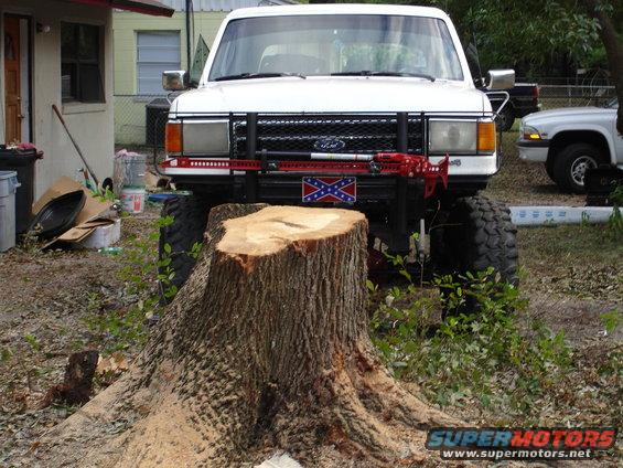 1989 Ford Bronco with Confederate flag and Hi-Lift jack, parked in front of a tree stump