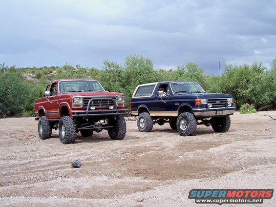 1987 Ford F-150 pickup and a blue Ford Bronco parked on dirt with scrub brush in background