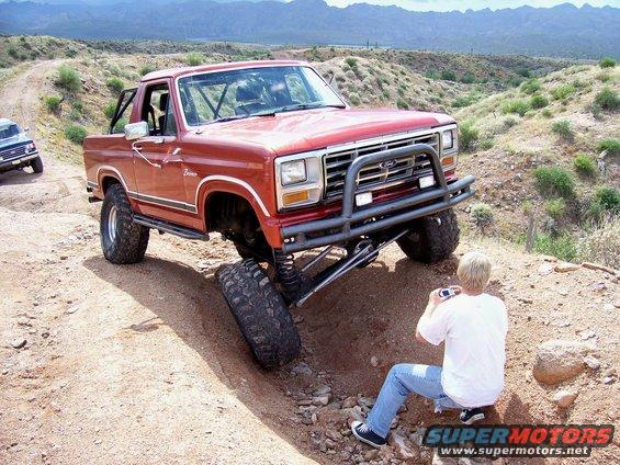 Red Ford Bronco with lifted suspension navigating rough terrain, person photographing