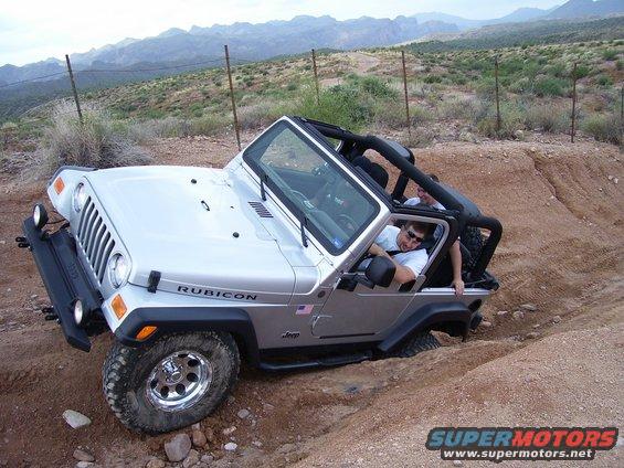 Silver Jeep Wrangler Rubicon driving on a dirt trail with two people inside. Mountains in background