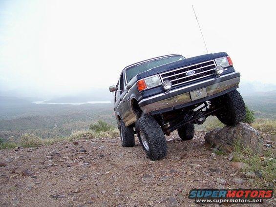 1991 Ford Bronco climbing a rock on a dirt hill with a lake in the background