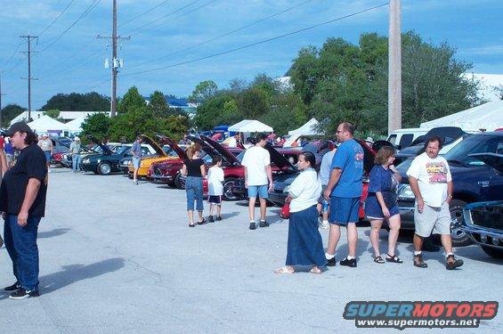 Outdoor car show with many vehicles and people milling around. Tents are visible in the background
