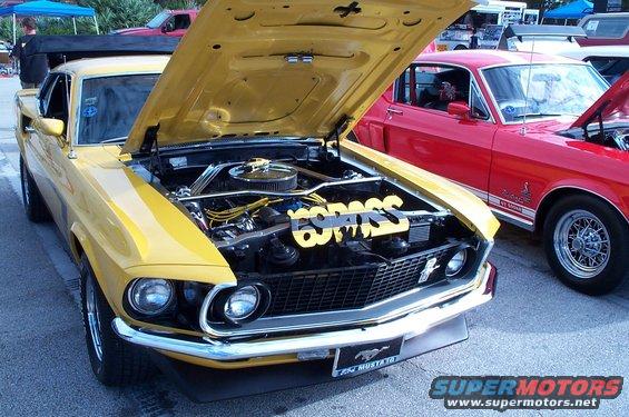 Yellow classic Ford Mustang with hood open, showing engine and a yellow "BOSS" decoration. Red classic Ford Mustang parked next to it