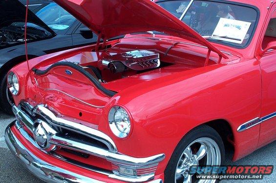 Red classic car with chrome grille and hood open, showing engine. Another black car is visible behind it