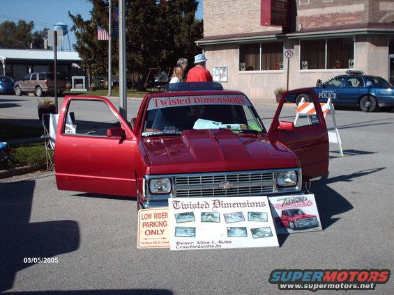 Red pickup truck with hood scoop and doors open, "Twisted Dimensions" banner on windshield