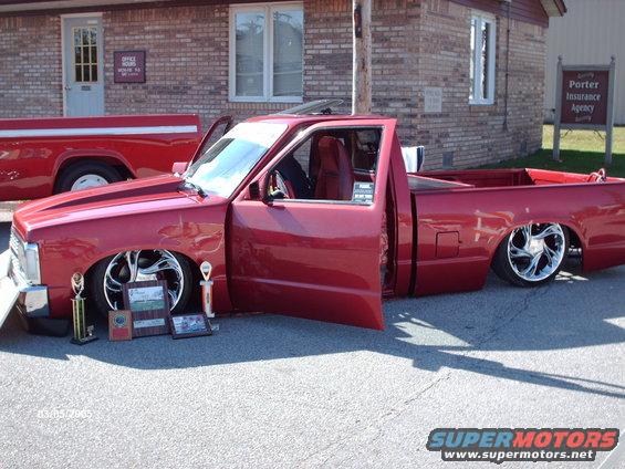 Red pickup truck with doors open, lowered suspension, and custom wheels. Trophies and awards are displayed in front