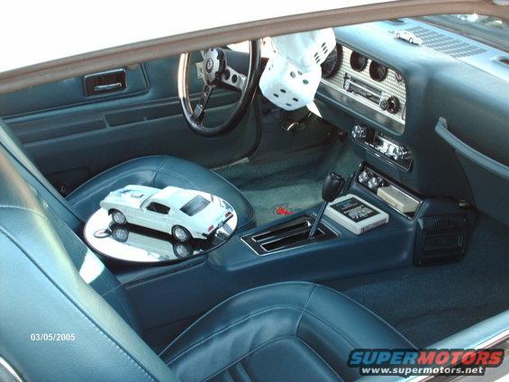 Interior view of a classic car, showing blue seats, dashboard with radio, and a white model car on the passenger seat. Dice hang from the rearview mirror