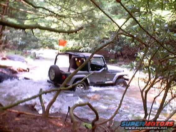 Gray Jeep Wrangler driving through a stream, spare tire mounted on the rear, orange flag on antenna