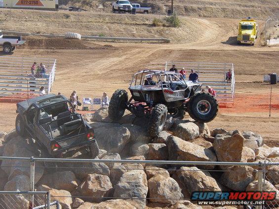 Two custom off-road vehicles navigating a rock obstacle course. One is a dark-colored, open-top vehicle, and the other is a silver buggy with a roll cage