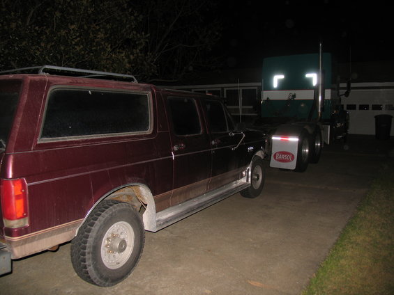 Maroon Ford Bronco SUV parked next to a green semi-truck at night. The SUV has a roof rack and a brown stripe along its side
