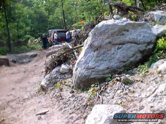 Red SUV and another vehicle stopped on a dirt path with large rocks and trees around