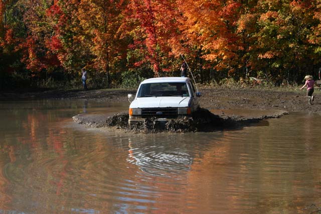 1987 Ford Bronco driving through a muddy puddle with fall foliage in the background