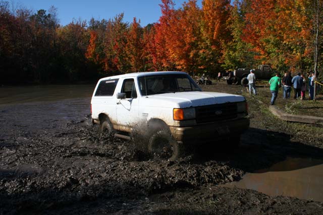 White SUV splashing through mud, trees with fall colors in background. Other vehicles and people visible