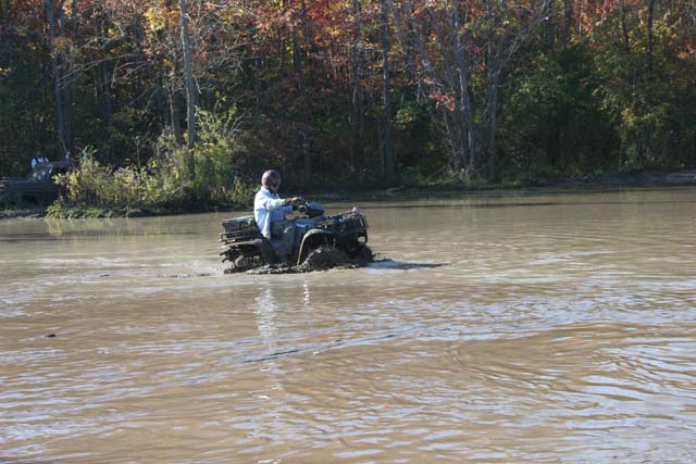 Person riding an ATV through muddy water with trees in the background. Another ATV is visible to the left