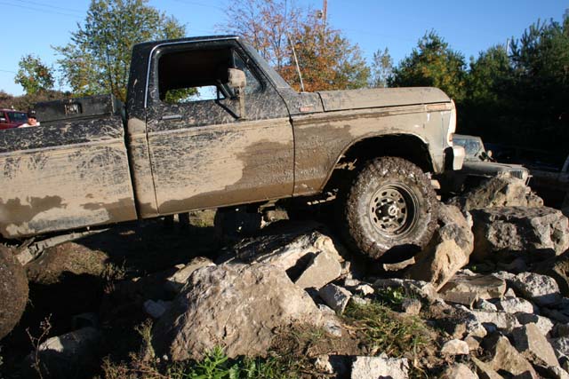 Mud-covered pickup truck with a toolbox on the bed, navigating large rocks. Another vehicle is partially visible behind it