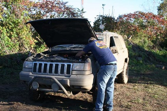 Man working on engine of muddy Jeep Grand Cherokee with hood up, off-road setting