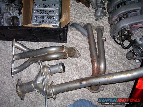 Car exhaust manifold and pipe parts laid out on carpet. Box with labels '93 MX6 LS MTX' visible