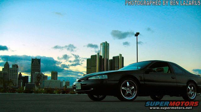 Black 1997 Ford Thunderbird coupe with chrome wheels parked in front of Detroit skyline