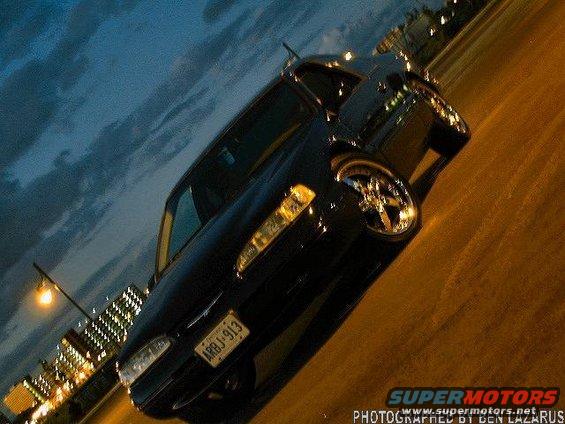 Black sedan with custom rims parked on a street at dusk, city lights in background
