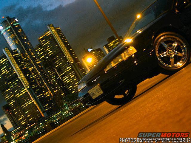 Black 1997 Ford Thunderbird with aftermarket wheels, seen from a 3/4 front angle at night with city lights in the background