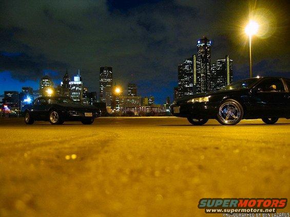 Two black cars parked at night, city skyline behind them
