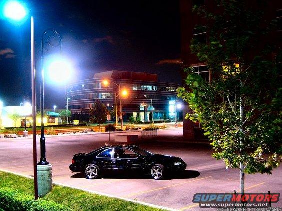 Black coupe parked in a lot at night, with a modern building in the background