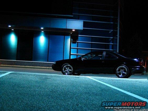 Black coupe parked at night, side view. Chrome wheels, tinted windows, modern building in background