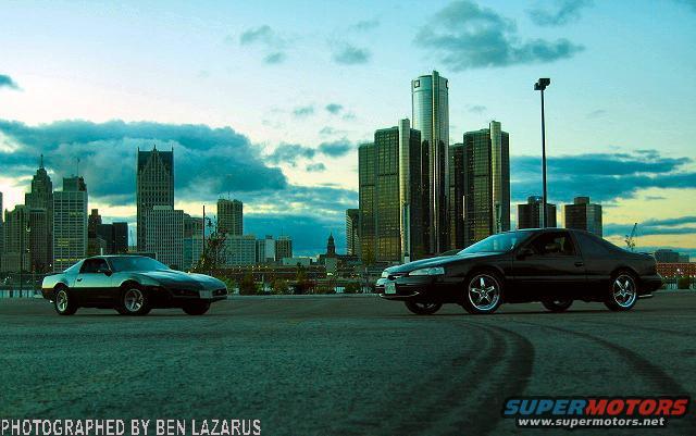 Black coupe and a lighter coupe parked with Detroit skyline behind them