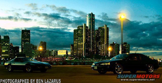 Two cars, a dark coupe and a black sedan, parked with a city skyline at dusk