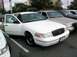 1998 Ford Crown Victoria - A white 1998 Ford Crown Victoria is parked with its driver's door ajar, alongside other vehicles.
