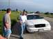 1998 Ford Crown Victoria - A white 1998 Ford Crown Victoria is parked on the side of a rural road with two men standing nearby.