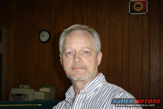 Man in striped shirt against wood-paneled wall with a clock and a logo