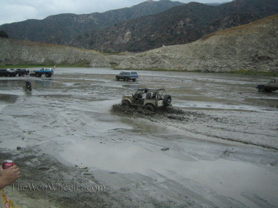 Jeep-like vehicle splashing through mud with other trucks and SUVs in the background near a lake and hills