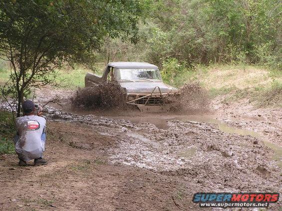 Dark gray 1978 Ford Bronco driving through mud, front bumper visible, splashing water