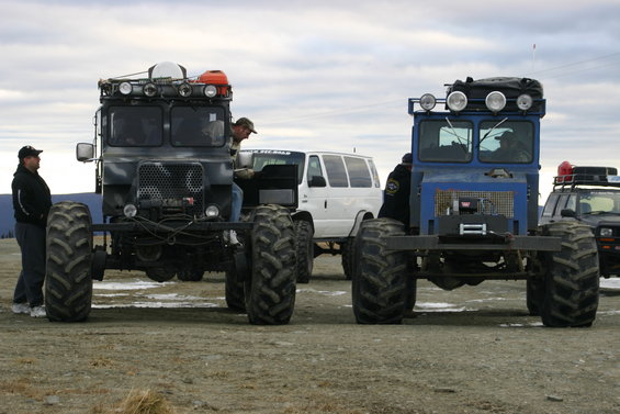 Two custom trucks with massive tires and roof racks, parked on dirt. A white van is behind them