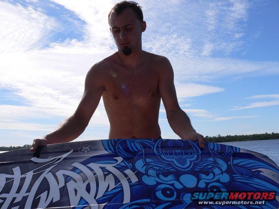 Man holding a blue and white wakeboard with dragon graphics. SUPERMOTORS.NET logo visible