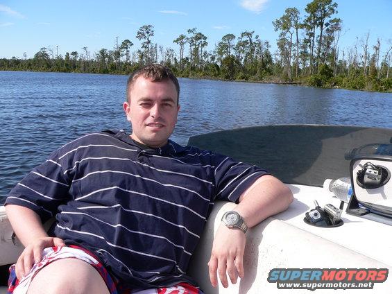 Guy in striped shirt and shorts on a boat, water and trees in background. Supermotors.net logo visible