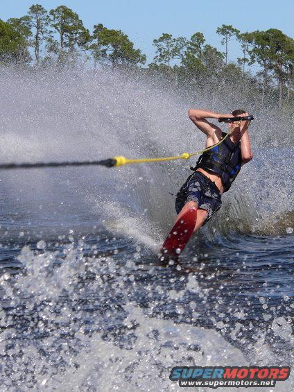 Waterskier in blue vest and patterned shorts on a red ski, holding a tow rope, with trees and blue sky in the background
