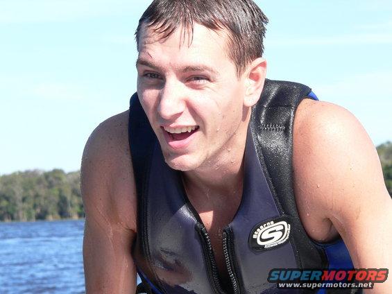 Man in blue life vest, smiling, on a body of water with trees in the background