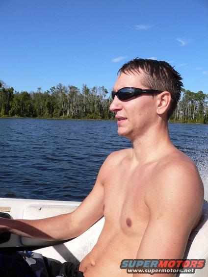Man wearing sunglasses in a boat on the water, trees in background