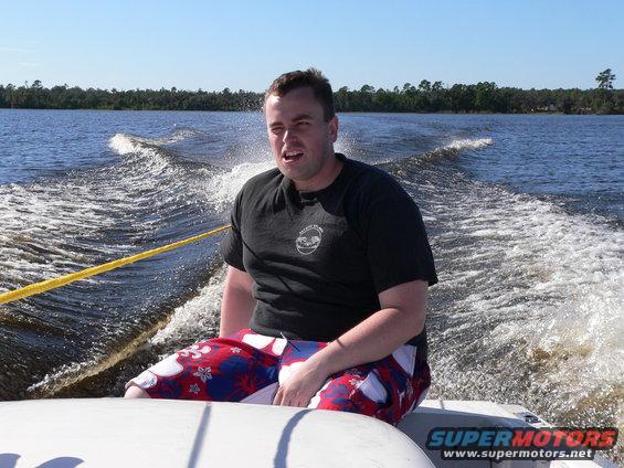 Person in a black t-shirt and patterned shorts sits on the edge of a white boat, being pulled through choppy water