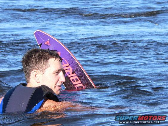Man in water wearing life vest, purple wakeboard behind him. Supermotors.net logo in corner