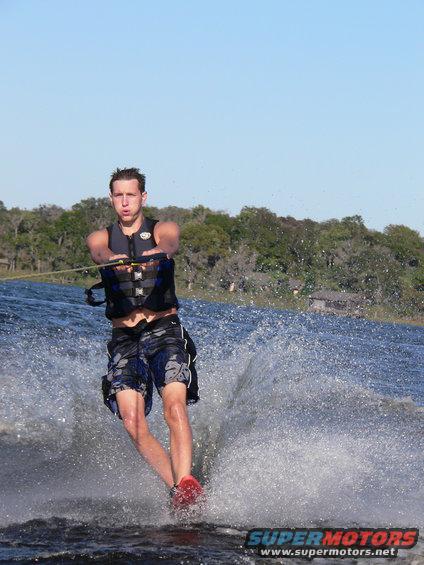 Man water skiing with a rope and handle, wearing a life vest and shorts. Water spraying