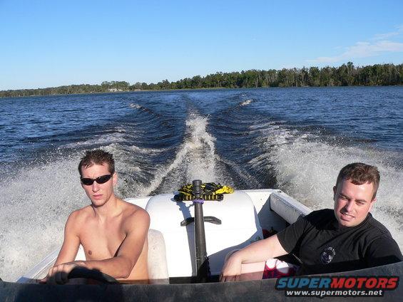 Two guys in a boat speeding across a lake, leaving a large wake. One is wearing sunglasses