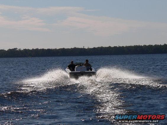 White boat with two people speeds across blue water, leaving a large wake