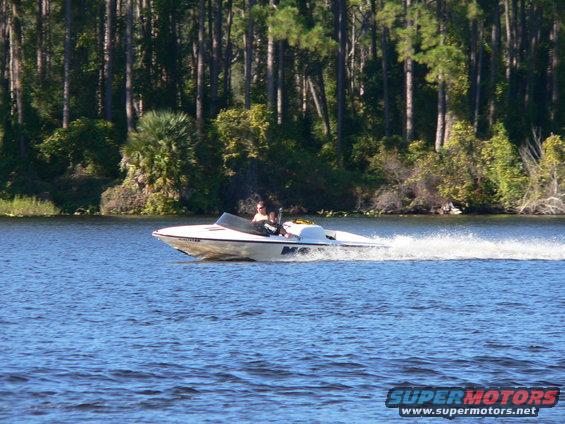 White speed boat with two people on board moving across blue water, trees in background