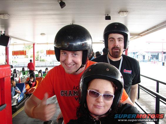 Three people wearing helmets at an amusement park, with go-karts visible in the background