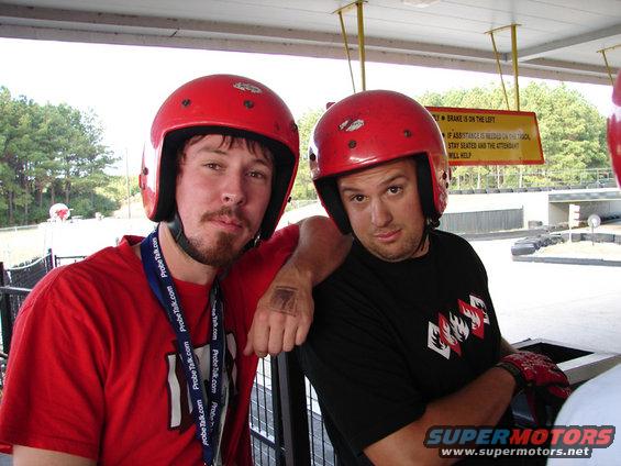 Two men in red helmets and racing attire at an outdoor track, one with a red shirt and lanyard, the other in a black shirt