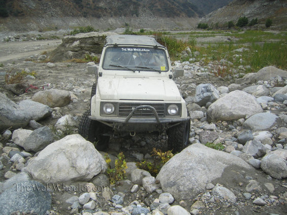 White 1987 Suzuki Samurai driving through a rocky stream bed