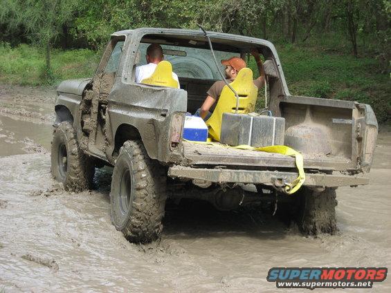 Muddy SUV with two people inside driving through a muddy trail. It has yellow seats and some gear in the back